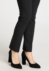 Decollete nere in suede con punta affilata e tacco largo, abbinate a pantaloni neri attillati, che mostrano una texture liscia.