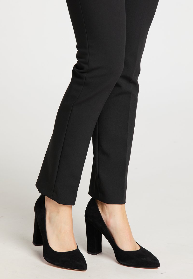 Decollete nere in suede con punta affilata e tacco largo, abbinate a pantaloni neri attillati, che mostrano una texture liscia.
