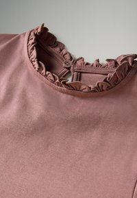 Detailný pohľad na výstrih fialovej košele s volánikovým lemom a jednotlivým zapínaním na gombík na svetlom pozadí.
