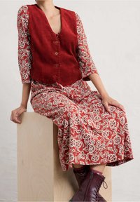 Gilet en velours côtelé rouge avec un col en V et des boutons superposé sur une robe à imprimé floral rouge et blanc, associée à des bottes à lacets bordeaux.