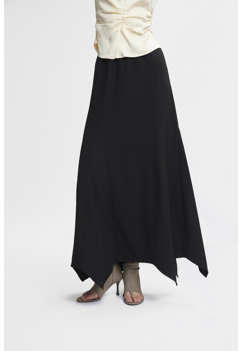 Gonna maxi nera con orlo asimmetrico, realizzata in un materiale fluido, con cuciture laterali e una texture liscia. Indossata con un top leggero.