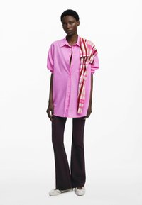 Roze oversized shirt met knoopsluiting, gecombineerd met donkere flared jeans. Een gebloemd sjaaltje hangt over één schouder. Sneakers maken de outfit af.