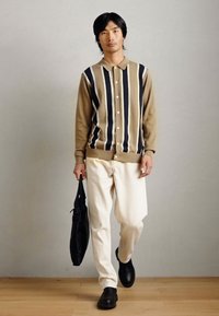 Beige och marinblå randig knapplåsvärmare, krämfärgad byxa, svarta skor, håller en svart väska, mot en neutral bakgrund.