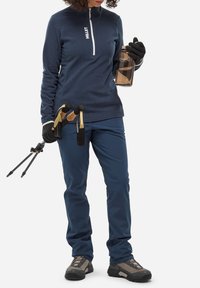 Marineblaue Fleece-Pullover mit Viertelreißverschluss, blaue Hose, schwarze Handschuhe, hält eine Wasserflasche und Trekkingstöcke. Wanderschuhe mit grauen Akzenten.