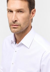 Weißes Hemd mit Kentkragen, das eine subtile Textur, Knopfverschlüsse und eine einzelne Brusttasche aufweist. Minimalistisches Design.