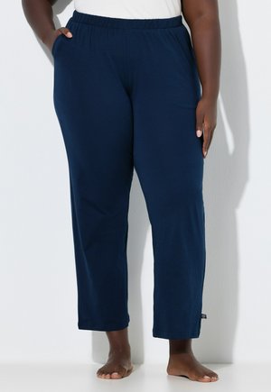 Persona con pantalones de cintura elástica azul marino, con las manos en los bolsillos, de pie descalza contra una pared blanca.