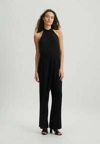 Čierny halter overal s voľnými širokými nohavicami, s viazaním v pase. Hladká látka s elegantným dizajnom. Skombinovaný s čiernymi sandálmi na podpätku.