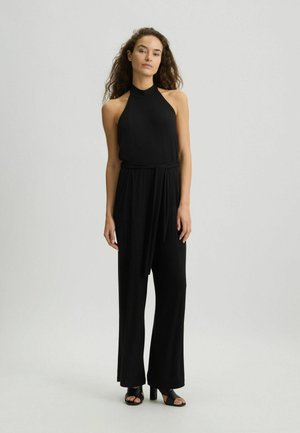 Zwarte halter jumpsuit met vloeiende wijde benen, voorzien van een strik in de taille. Glad materiaal met een strakke uitstraling. Gecombineerd met zwarte hakken.