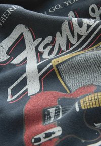 Červená grafika elektrickej gitary s zosilňovačom a veľkým štýlizovaným nápisom „Fender“ na tmavom látkovom pozadí.