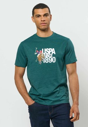 Homme portant un t-shirt graphique vert USPA avec un motif joueur de polo et un jean bleu foncé, debout avec une main dans la poche.