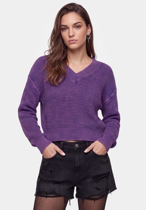 Jonge vrouw met lang bruin haar, draag een gestructureerde paarse trui en zwarte gescheurde denimshorts met handen in de zakken.