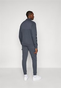 Mørk grå treningsdrakt med en lysere grå overdel, som har subtil merking på ermene. Slim fit med ribbestrikkede mansjetter og lommer.