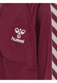Vinröd sweatshirt med dragkedja, prydd med en vit "hummel"-logotyp. Ärmen har kontrasterande diagonala randiga detaljer i vitt. Mjuk textur.