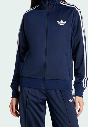 Person in einem marineblauen Adidas-Trainingsanzug mit weißen Streifen an Ärmeln und Hose, das Adidas-Trefoil-Logo ist auf Brust und Oberschenkel sichtbar.
