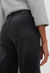 Jean en denim noir avec une texture lisse, taille mi-haute et deux poches arrière. Le modèle porte un pull gris clair.