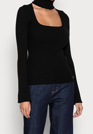 Mujer con camisa negra de manga larga acanalada con escote cuadrado y cuello alto recortado, combinada con vaqueros de mezclilla azul oscuro.
