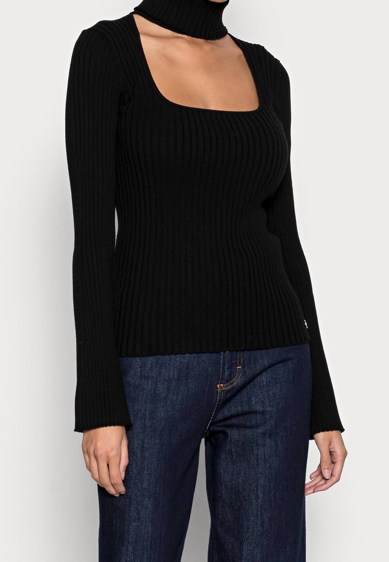 Femme portant un haut noir côtelé à manches longues avec un décolleté carré et un col roulé découpé, associé à un jean denim bleu foncé.
