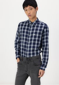 Navy and gray plaid shirt with long sleeves and button closure, made of soft fabric; worn with dark gray jeans. Small logo on the chest.