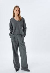 Grijze V-hals trui met een losse pasvorm, gecombineerd met grijze broek met wijde pijpen. De broek heeft plooien en loopt taps toe bij de enkels. Geweven stof.