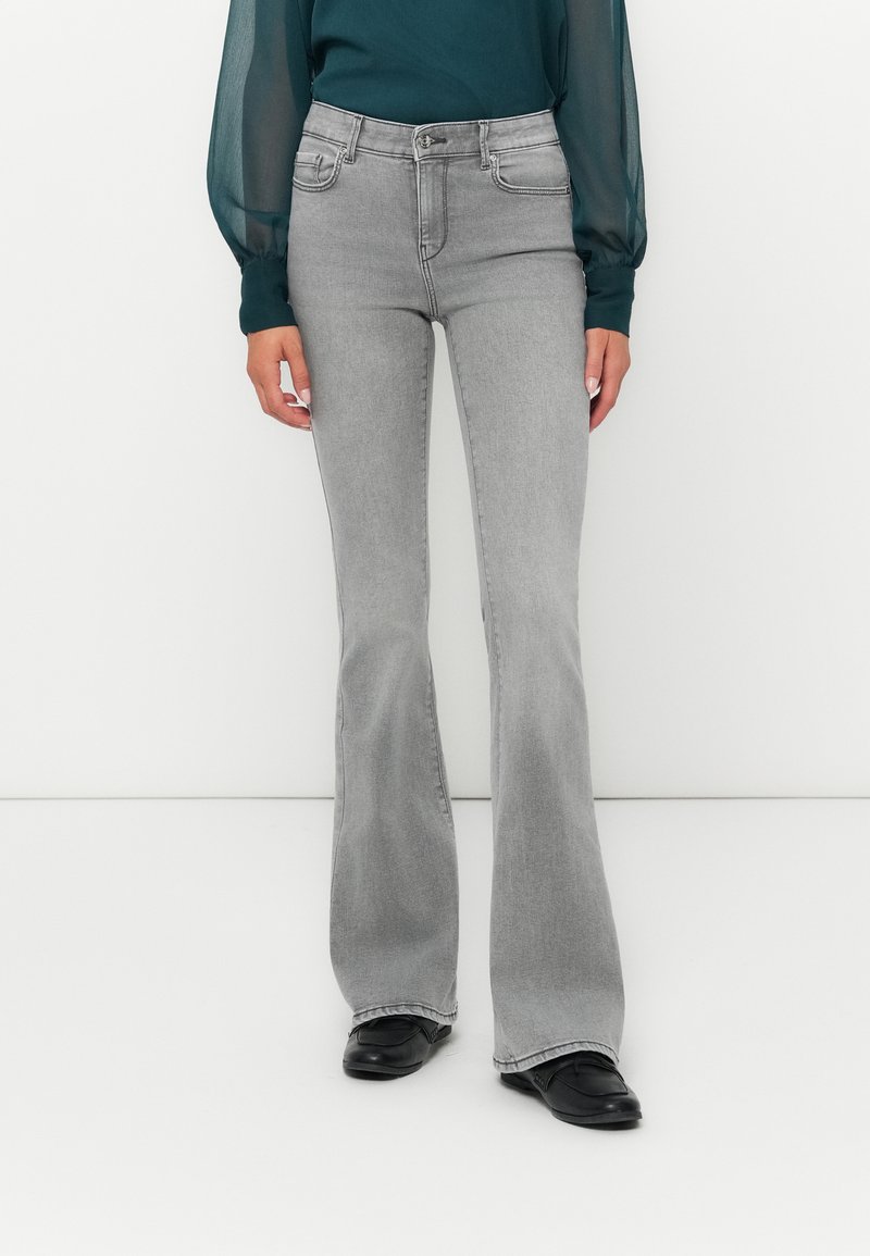 Jeans grigi flared con vita alta, dotati di tasche anteriori e una texture liscia. Abbinati a un top lungo a maniche trasparenti di colore teal.