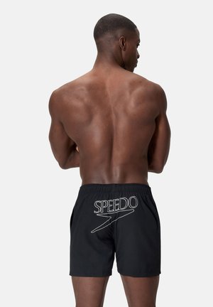 Uomo muscoloso di schiena, indossa un costume da bagno Speedo nero con logo bianco sul retro, su uno sfondo chiaro e uniforme.