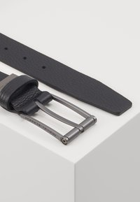 Correa de reloj de cuero negro con un acabado texturizado, que cuenta con una hebilla de metal gris mate y un logo. La correa es estrecha y tiene extremos planos.