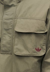 Ζακέτα χακί με φερμουάρ, κατασκευασμένη από λείο ύφασμα, διαθέτει εμπρόσθια τσέπη και κεντημένο λογότυπο μπορντό.