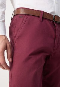 Burgunderfarbene Hose aus glattem Stoff, mit einem braunen Ledergürtel. Dekorative Nähte und ein kleines Logo-Detail am Bund.