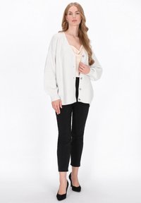 Jasnoszary sweter dzianinowy z balonowymi rękawami, z dekoltem w kształcie litery V i srebrnymi guzikami, noszony na kremowej bluzce na ramiączkach i zestawiony z czarnymi spodniami.