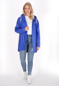 Zila ūdensizturīga lietus jaka ar kapuci, regulējamiem sprādzēm, spiedpogām un taisnu griezumu, saskaņota ar gaišiem džinsiem un baltām sporta kurpēm.