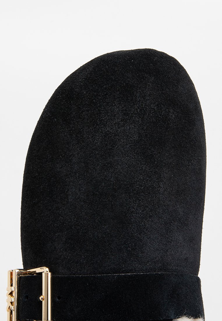 Bout de botte en daim noir avec une boucle en métal doré et un détail de sangle sur un fond blanc.