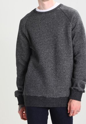 Man draagt een donkergrijze, gestructureerde trui met ronde hals over een wit hemd en donkere broek, staand tegen een effen lichte achtergrond.