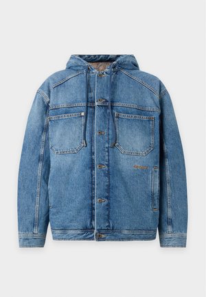 Joven con chaqueta vaquera azul sobre camisa blanca y camiseta negra, pantalones cargo beige con logotipos y zapatillas negras y blancas, de pie con la mano en el bolsillo.