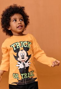 Orange langærmet T-shirt med en sort og grøn Halloween-grafik, der viser en tegneseriefigur, teksten "TRICK OR TREAT" og blødt stof.