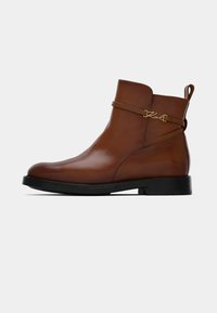 Brown leather ankle boot with a glossy finish, low black rubber sole, side strap detailing, and a gold logo accent.