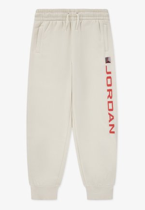 Beige jogger pants with elastic cuffs and waistband, side pockets, drawstring, and red "JORDAN" text with logo on one leg.