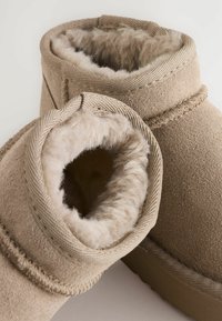 Beige ruskind ankelstøvler med fluffy uldforing og syet kant, vist i nærbillede mod en neutral baggrund.