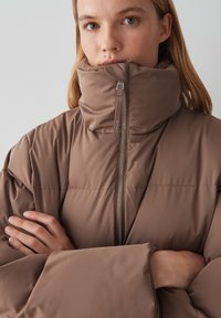 Giubbotto imbottito marrone con colletto alto, caratterizzato da una zip frontale e design trapuntato. Il tessuto appare liscio con una finitura leggermente lucida.