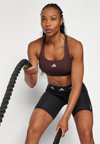 Brun sports-bh med hvit logo, kombinert med svarte shorts som har et midjebånd med logo. Bh-en har firkantet utrigning; tauet er grepet fast.