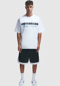 Tricou alb cu text negru și aqua care spune "MAI PUȚIN DERULAT MAI MULT TRAIT," asortat cu pantaloni scurți negri cu accente albe și pantofi de baschet.