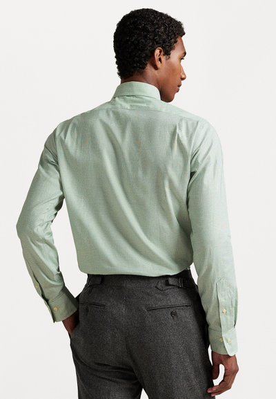 Bărbat întors cu spatele, purtând o cămașă de dress de culoare verde deschis, cu carouri mici, băgată în pantaloni de costum gri închis, cu mâinile în buzunare.