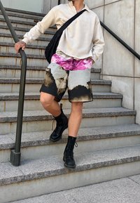Sudadera oversized de beige claro, pantalones cortos estampados en rosa y negro, y botas negras con cordones, de pie en unas escaleras con un bolso negro de hombro.