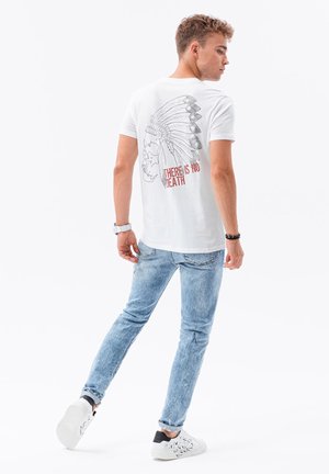 Weißes Baumwoll-T-Shirt mit einem schwarzen Grafikdesign eines Schädels und Federschmucks auf dem Rücken, mit rotem Text "ES GIBT DEN TOD NICHT." Getragen mit blauen Jeans.