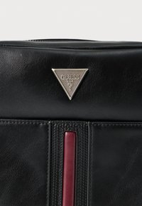 Fekete bőr táska, amelyen egy texturált, függőleges piros csík és egy háromszög alakú ezüst logó díszítés található. Simább felület, részletes varrásokkal.