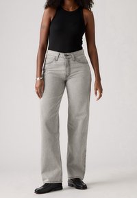 Femme portant un haut noir sans manches, un jean droit taille haute gris clair, des mocassins noirs et des bracelets argentés sur un fond neutre.