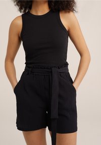 Žena v bezramínkovém černém žebrovaném topu a černých strukturovaných šortkách s vysokým pasem a uvázaným páskem, ruce má v kapsách.