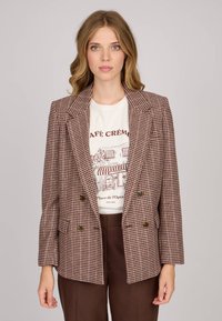 Blazer croisé en motif vichy marron et crème, avec larges revers, deux boutons et poches latérales. Porté par-dessus un t-shirt graphique.