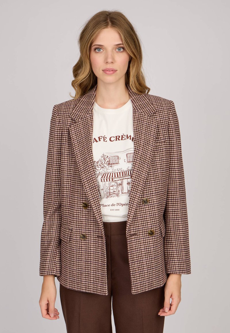 Blazer croisé en motif vichy marron et crème, avec larges revers, deux boutons et poches latérales. Porté par-dessus un t-shirt graphique.