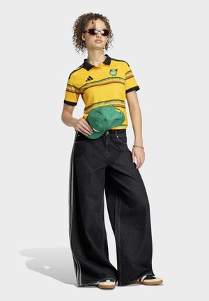 Junge Person trägt ein gelb gemustertes Poloshirt, schwarze Jeans mit weitem Bein und seitlichen Streifen, Sonnenbrille und hält eine grüne Baseballkappe.