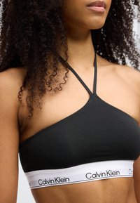 Μαύρο σουτιέν bralette με τριγωνικό σχεδιασμό, λεπτές τιράντες και λευκή ταινία με το λογότυπο "Calvin Klein" κατά μήκος του κάτω μέρους. Λεία υφή.